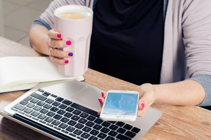 A woman, holding a cup of coffee and a cell phone, plus a laptop